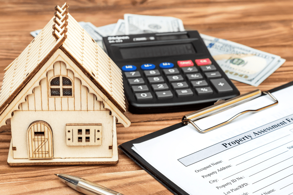 A small fake wood house, black handheld calculator, cash, and a clipboard with documents on top of a wood table.