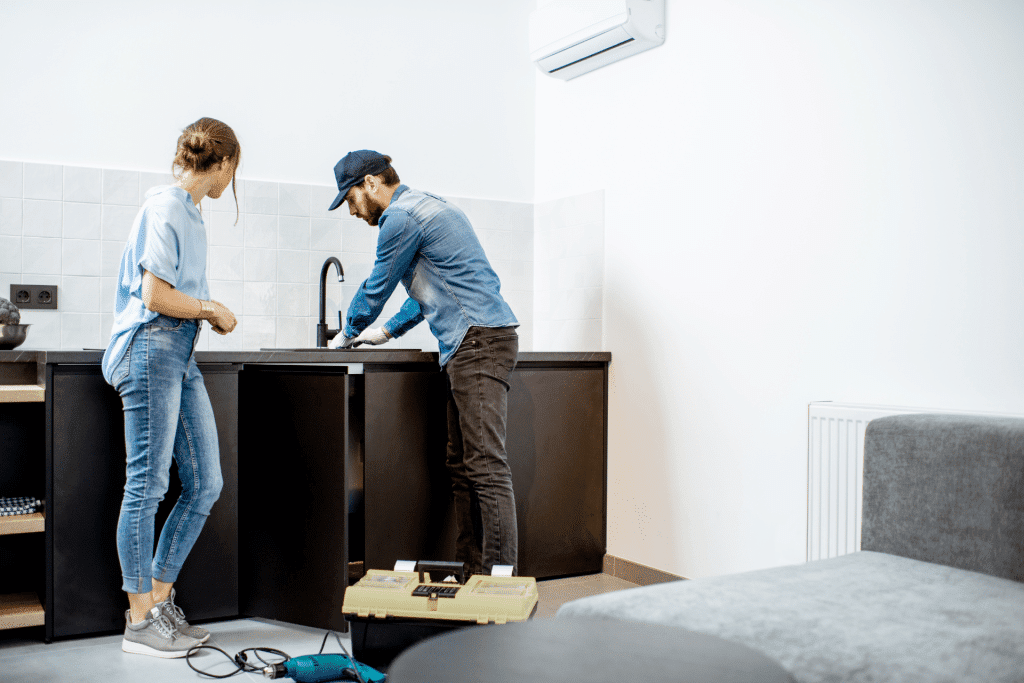 A maintenance man repairing a plumbing issue under a kitchen sink. I woman is standing next to him watching what he is doing.