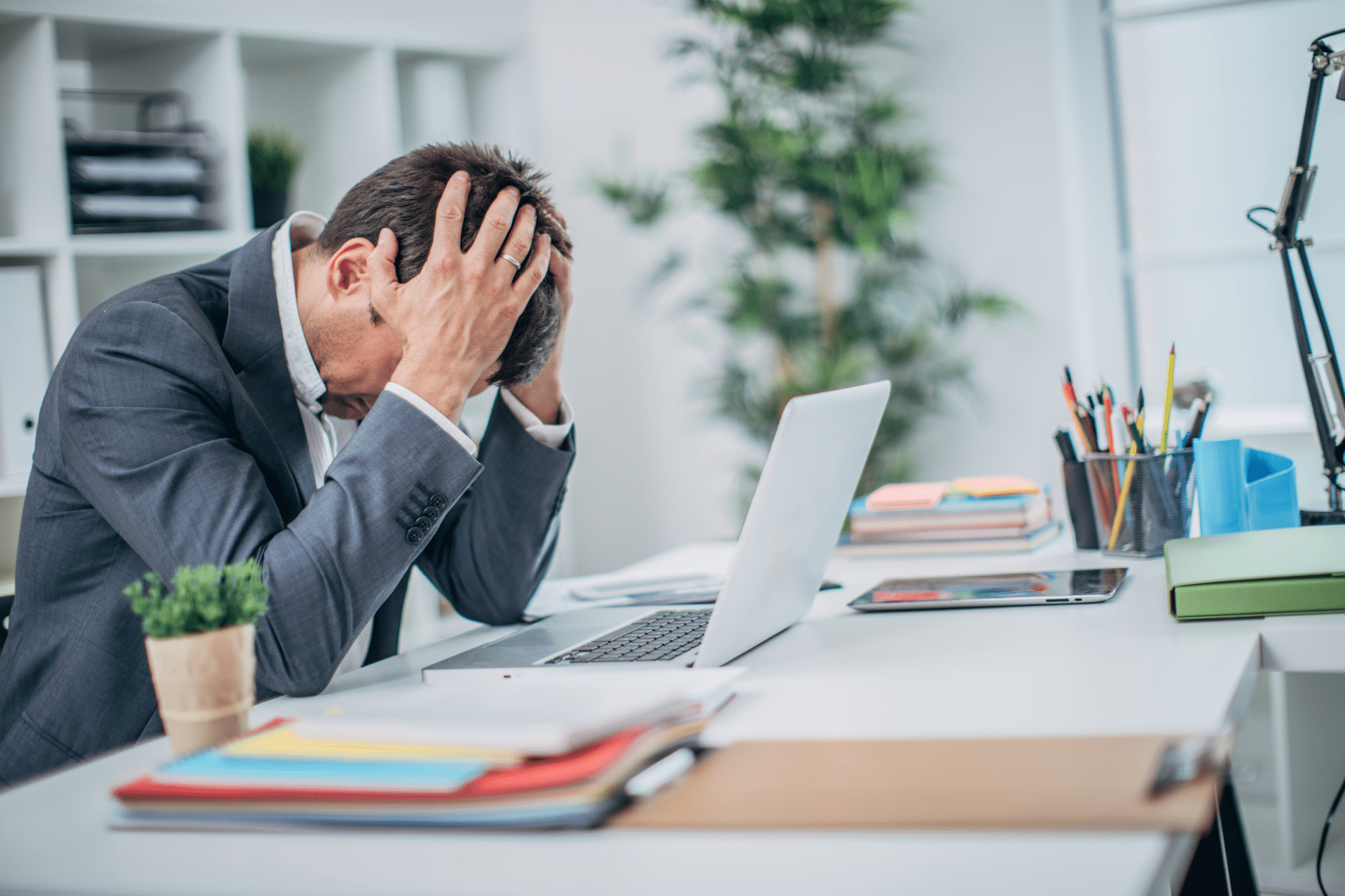 A man in a suit hunched over his desk in front of his laptop with his hands on his head. He looks distressed.