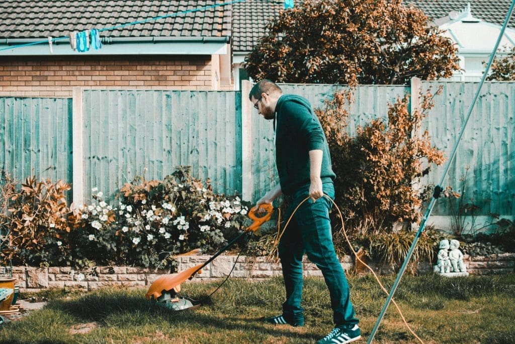 A man in a black hoodie, jeans, and shoes mows the lawn with an orange lawnmower in a backyard with a blue fence and white flowers, showcasing one of the summer property maintenance tips.