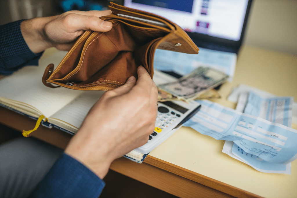 A man opening his empty wallet sitting in front of a desk. The desk has a computer, money, and check slips on it.