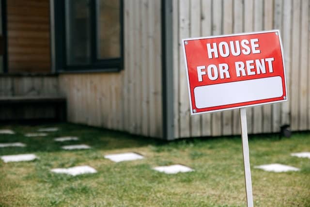 A red sign with the words "house for rent" on it, sticking into the ground, in front of a house.