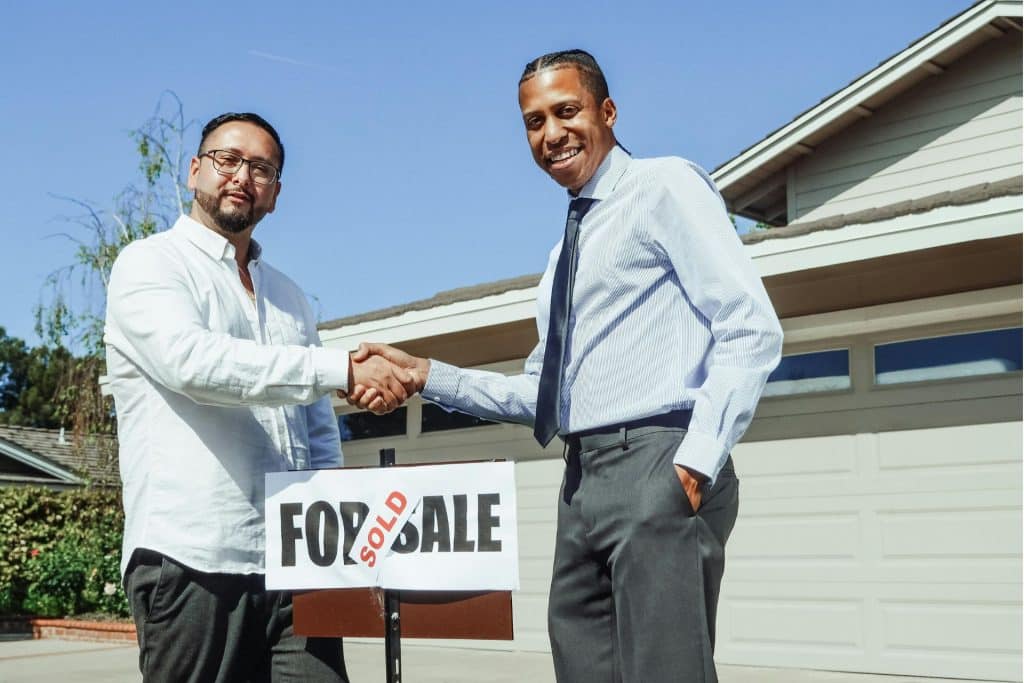 Real estate investing success is depicted as two businessmen in dress shirts shake hands in front of a white house with a 'Sold' sticker on the 'For Sale' sign between them