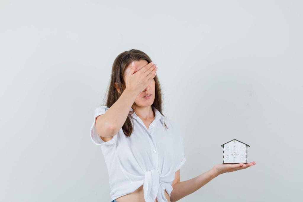 A frustrated woman with brown hair, wearing a white blouse, holding a miniature house model in her left hand, and covering her eyes with her right hand due to poor property management.