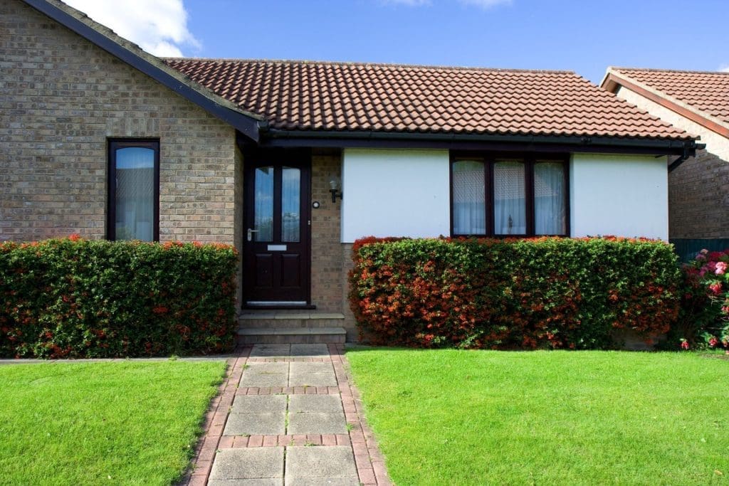 A brick house with a brown door and a brick pathway leads to a green lawn with red flower bushes, highlighting the curb's appeal, an essential property management tip for boosting rental value.