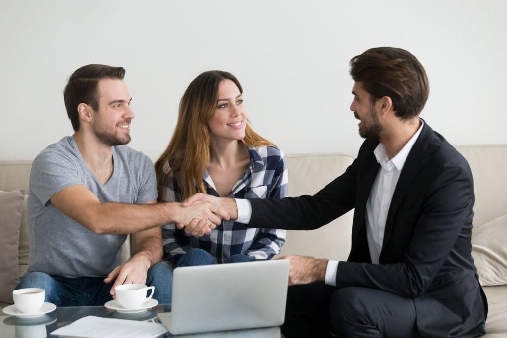 A man in a suit shakes hands with a smiling couple on a couch, symbolizing an agreement for finding good renters.