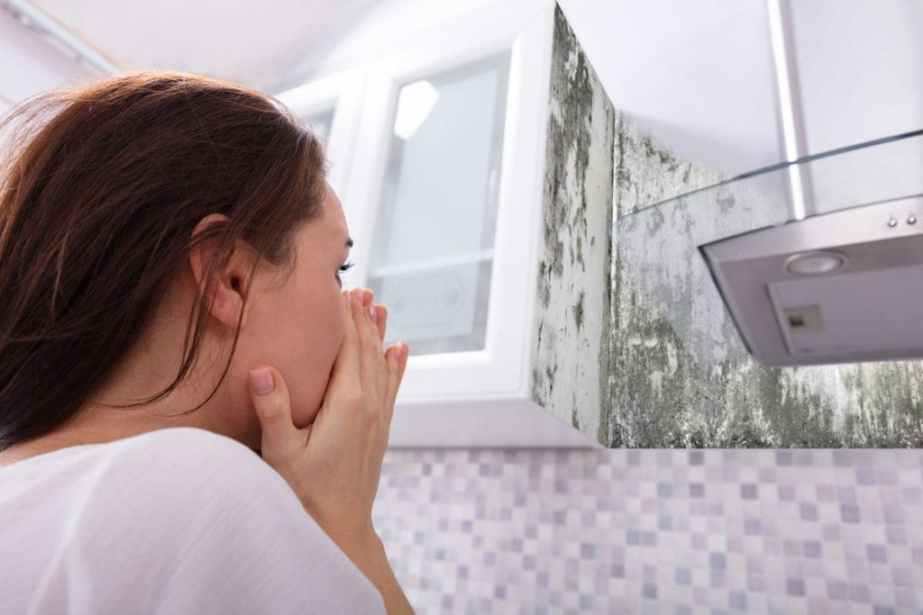 A woman looking in horror at mold growing on the wall of her kitchen, covering her mouth. The vent hood above highlights the mold at your rental.